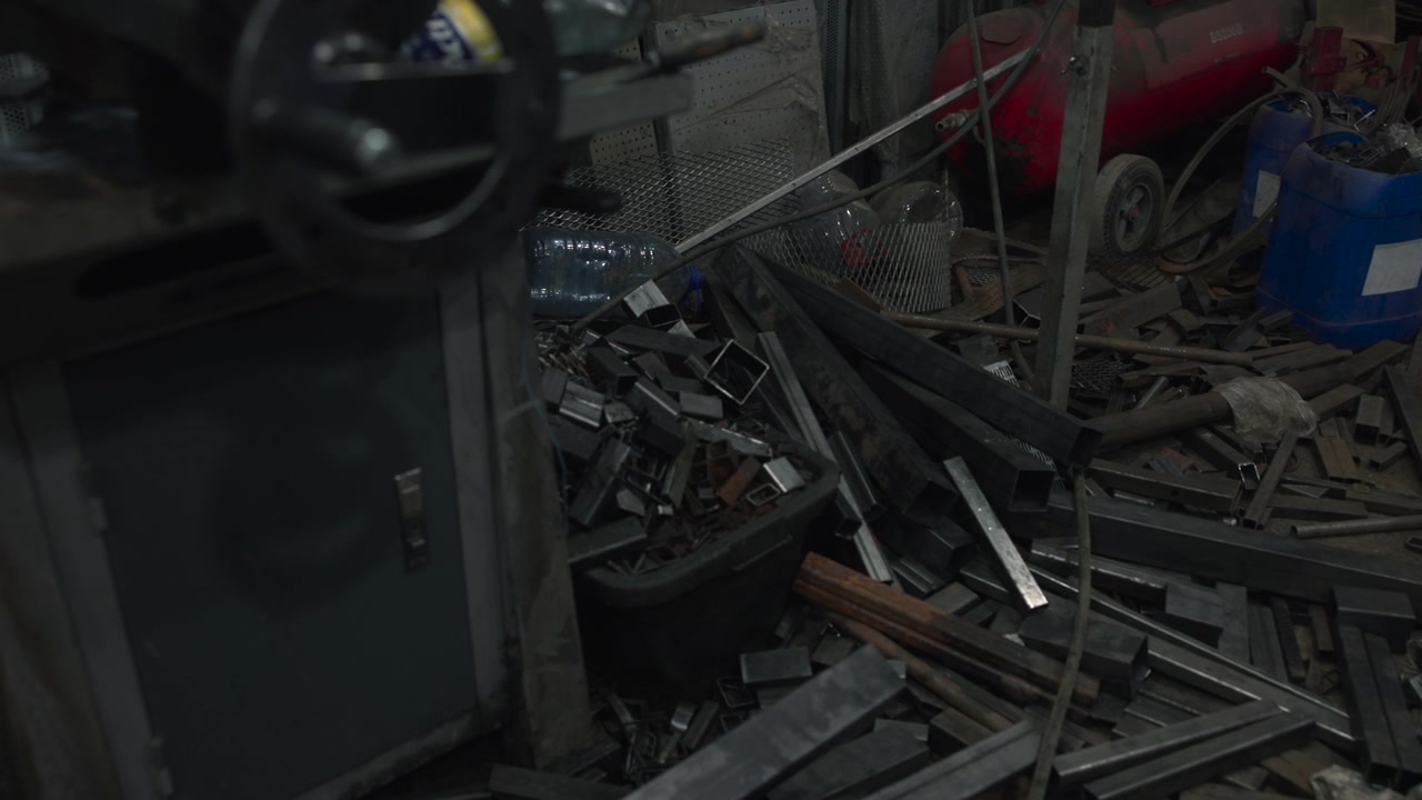 L'interno fioco di uno spazio di fabbricazione ingombro mostra rottami metallici sparsi, pannelli a rete, bombole di gas, cavi e stoccaggio grezzo sotto una luce bassa, creando un'atmosfera industriale cruda e una texture profonda