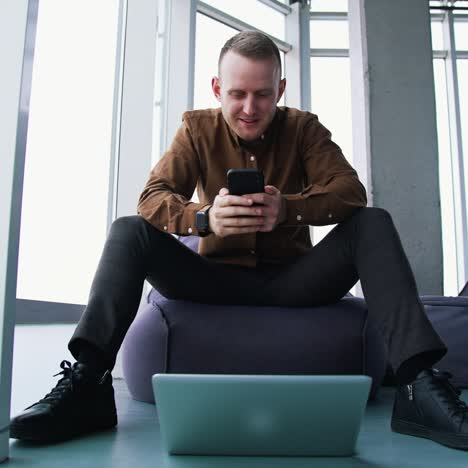 young man with a phone in office. handsome businessman sitting on a comfortable armchair and using a mobile phone on window background. front view