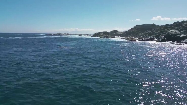 Volo planato basso sopra un oceano blu profondo scintillante che si avvicina a un promontorio roccioso spettacolare con ville costiere sotto un cielo limpido e luminoso