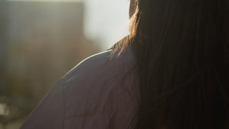 close-up of a girl with long brown hair flowing, wearing a peach jacket, walking through an urban area at sunset. the sunlight softly illuminates the side of her face