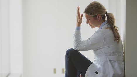 Stressed-Female-Doctor-Wearing-White-Coat-Sitting-On-Floor-In-Hospital-Corridor-With-Head-In-Hands