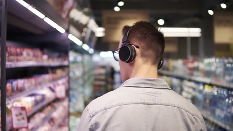 en el supermercado: un elegante tipo caucásico con auriculares camina a través de la sección de productos cárnicos de la tienda. siguiente toma de vista posterior. cámara lenta. fondo borroso