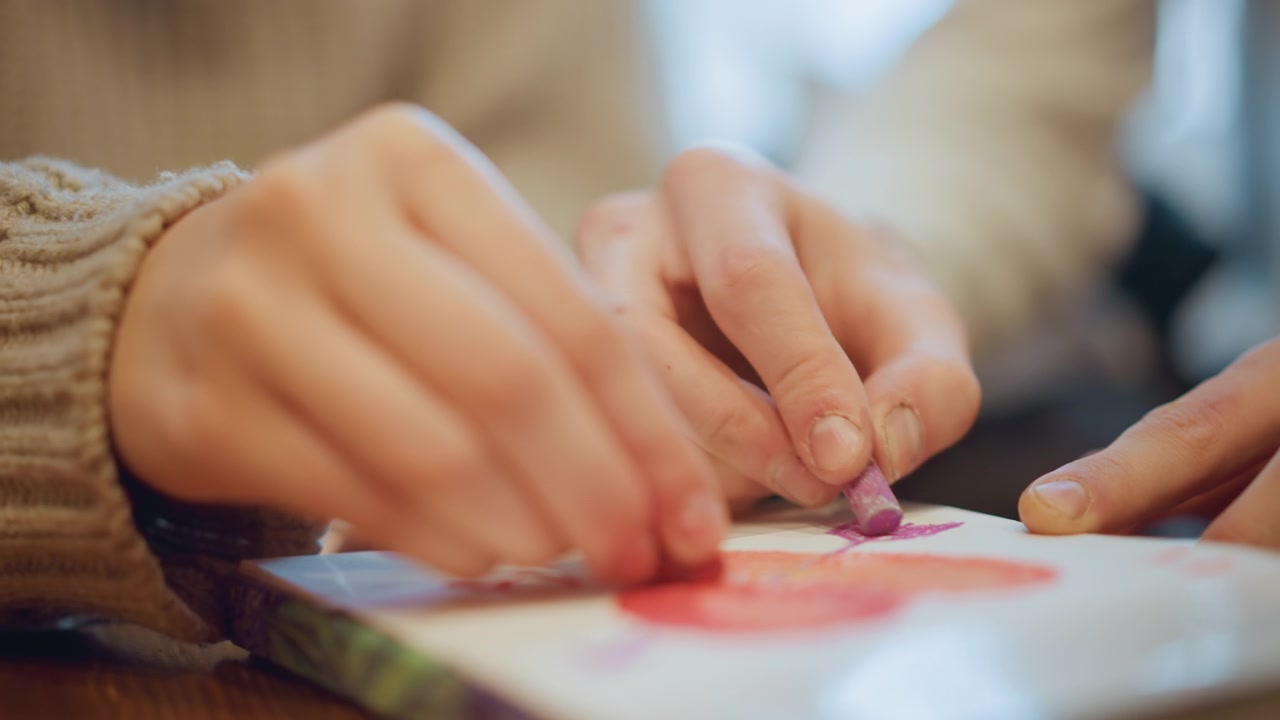 Primo piano di mani che disegnano con un pastello viola su carta bianca, creando un'opera d'arte colorata. La persona indossa una manica a maglia beige e la scena è caratterizzata da una luce interna soffusa in un ambiente di lavoro artistico.