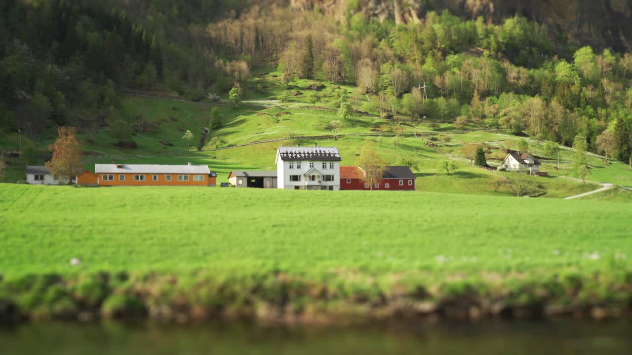 A farm near the Steinsdalsfossen waterfall, Norway. Three buildings sitting at the foot of the mountain, surrounded by the forest and the green farm fields. The river is slowly flowing by