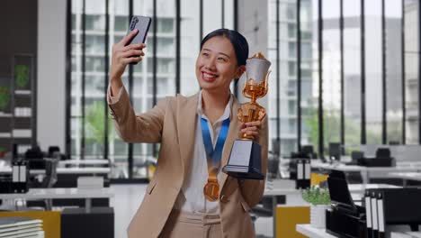 Asian Business Woman In A Suit With A Gold Medal Holding A Gold Trophy And Taking Photo On Smartphone As The First Winner In The Office