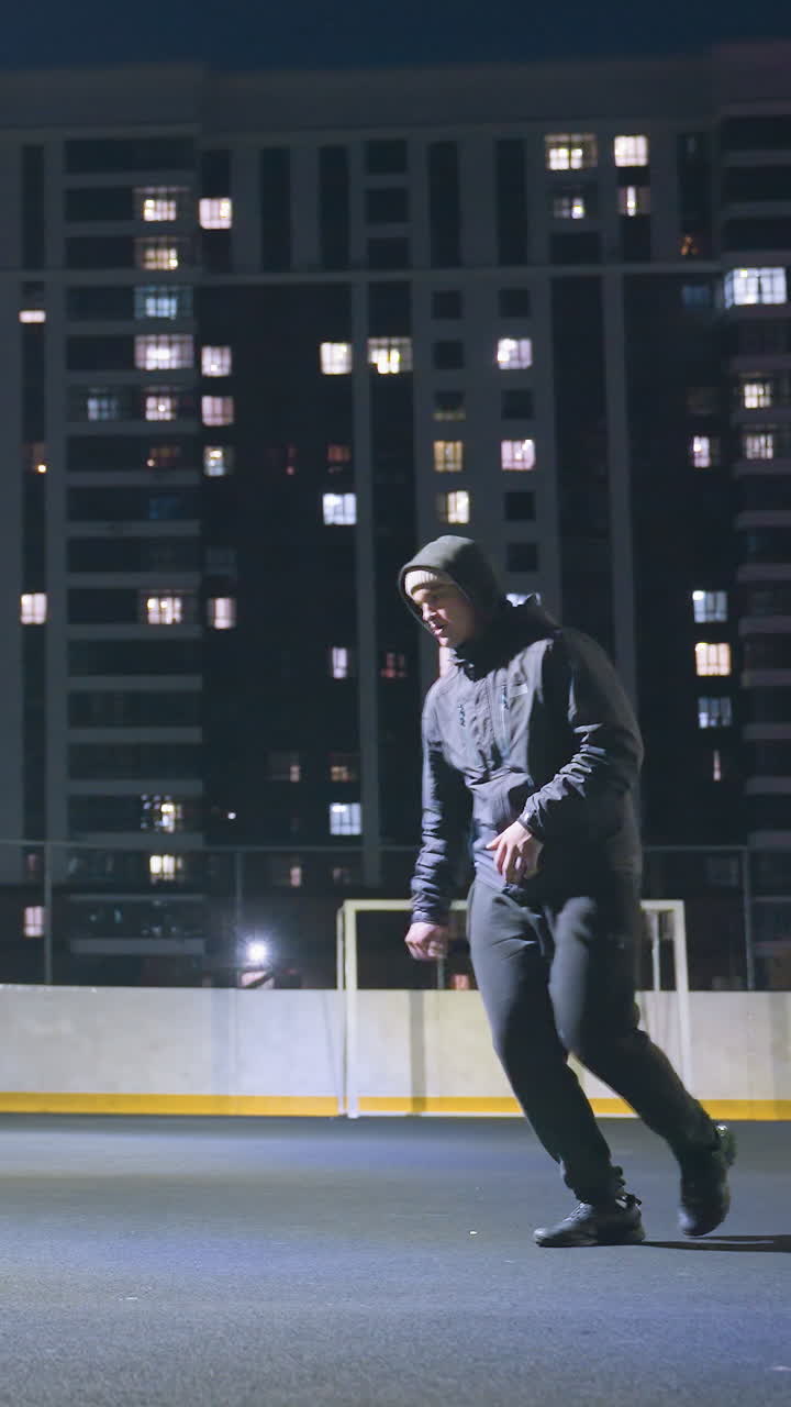 Side view of man in hoodie playing soccer under bright stadium lighting at night, ball near goalpost with reflection on wall, surrounded by urban cityscape and illuminated office buildings