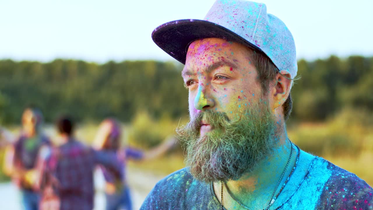 el primer plano del joven apuesto con barba y sombrero girando la cara hacia la cámara y sonriendo mientras estaba en manchas coloridas de pinturas mientras celebraba el festival holi con amigos