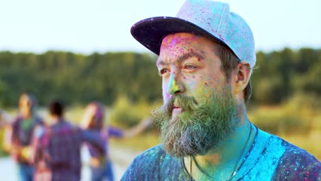 close up of the young handsome man with a beard and in a hat turning face tothe camera and smiling while being in colorful spots of paints as celebrating holi festival with friends