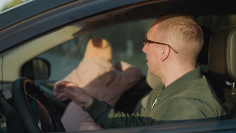 a man in a green jacket sits in a car, adjusting his glasses while a young girl in a pink jacket and knit hat looks out the window