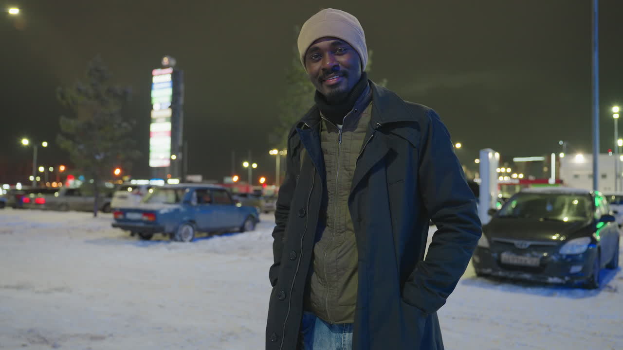 Man in winter coat and hat stands outdoors on snowy night with parked cars and illuminated city lights in background, posing with relaxed expression in cold season urban environment