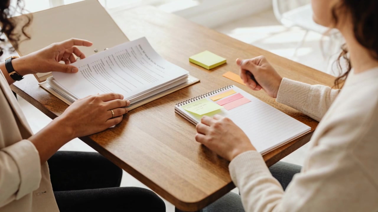 Flipping binder, left woman in blazer handing sheet at wooden table for review with sticky notes