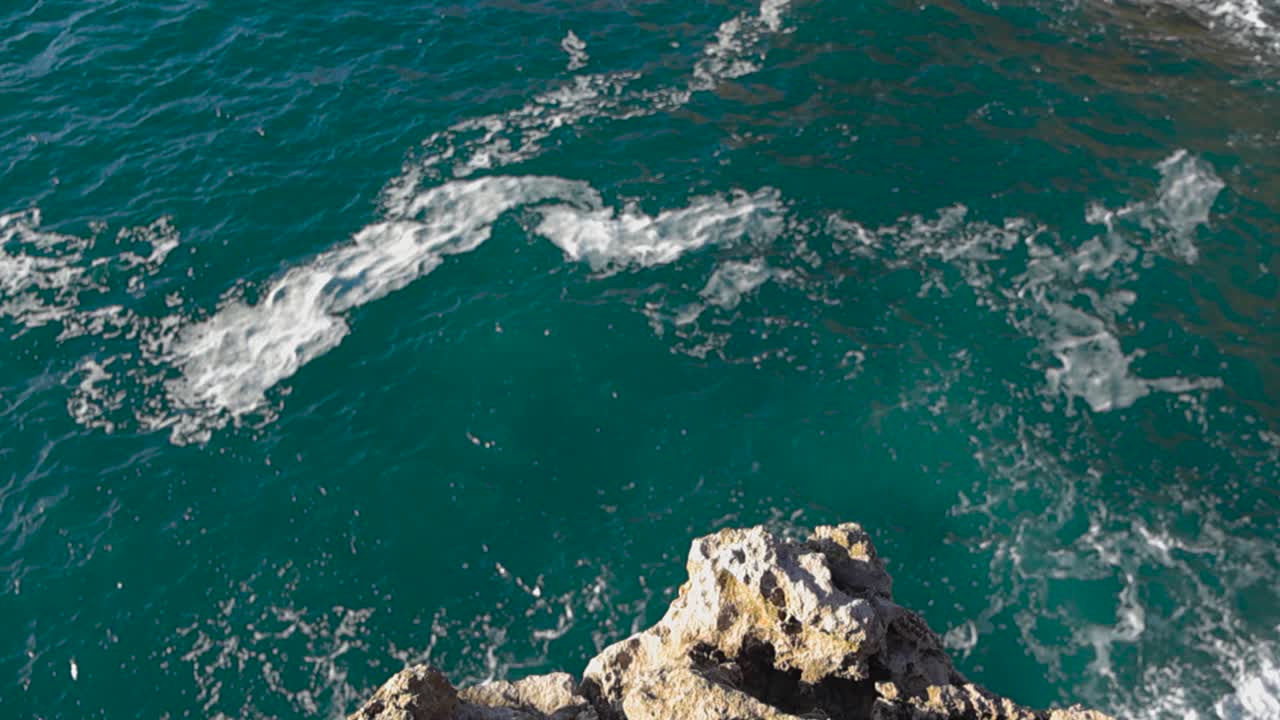 Lenta panoramica discendente dall'alto di scogliere costiere che rivela le acque turchesi e vorticose dell'Atlantico sottostante, con correnti spumeggianti e un movimento potente che crea una vista marittima spettacolare e ipnotica con onde oceaniche