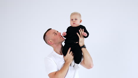 proud caucasian dad in white t-shirt is holding a baby on his shoulder. father and son portrait at white backdrop.