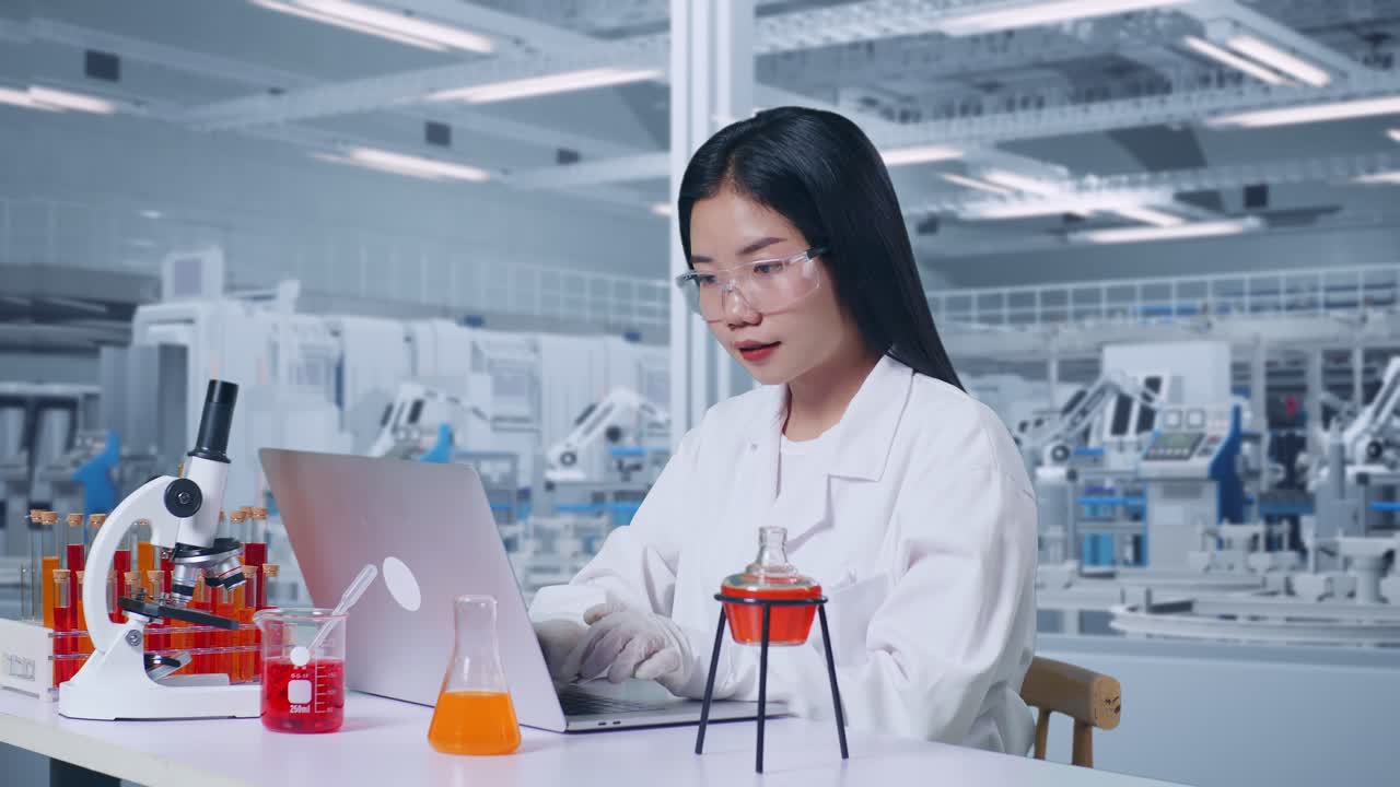 아시아 여성 과학자가 실험실에서 현미경과 시험관으로 테이블에서 작업하는 동안 노트북으로 타이핑하는 측면 뷰.
