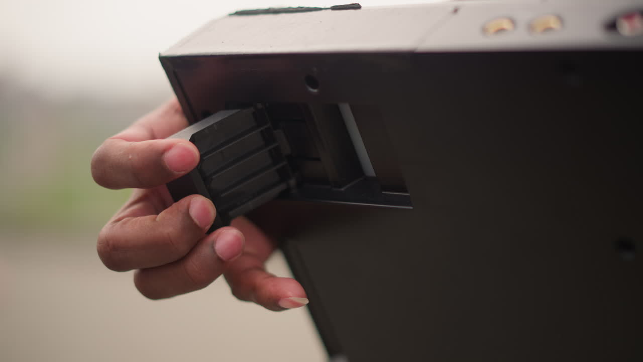 Closeup hand inserting battery module into slot, deliberate fingers sliding multicell pack into rear compartment of compact controller, urban bokeh background, tactile touch, subtle anticipation