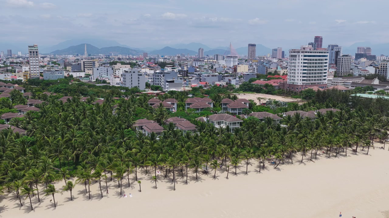Cảnh quay drone thiết lập ban ngày từ trên cao bãi biển cát với những cây cọ, biệt thự nghỉ dưỡng và đường chân trời thành phố ở Đà Nẵng Việt Nam.
