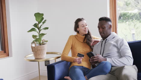 happy diverse couple sitting on sofa, using smartphone and credit card in home,copy space