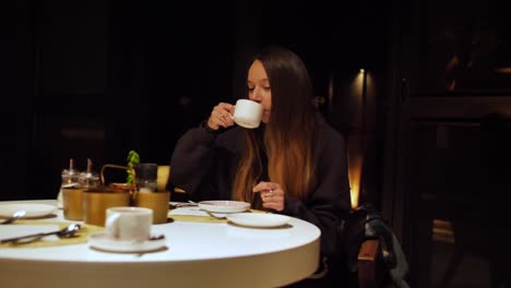 Toma-En-Cámara-Lenta-De-Una-Mujer-Joven-Sentada-En-La-Oscuridad-En-La-Mesa-Del-Desayuno-Bebiendo-Su-Café