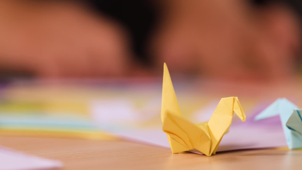 Una gru di carta gialla è appoggiata su un tavolo di legno, mentre delle mani piegano della carta sullo sfondo