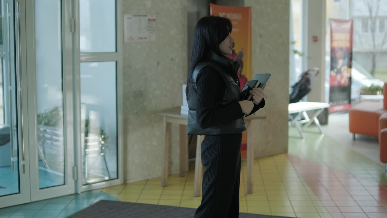 gerente femenina profesional en un traje formal negro, caminando por la oficina, interactuando con colegas, sosteniendo documentos y una computadora portátil, mostrando liderazgo y profesionalismo