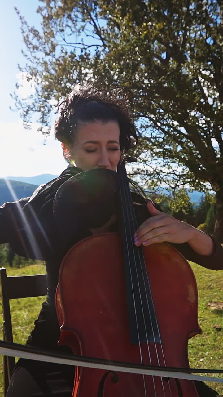 Emotional female musician playing bass-viol in the mountains. Lady in dress sits on the chair playing instrument at the backdrop of beautiful landscape. Vertical video