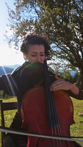 emotional female musician playing bass-viol in the mountains. lady in dress sits on the chair playing instrument at the backdrop of beautiful landscape. vertical video