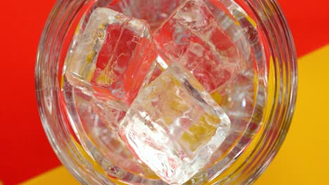 vista de pájaro disparo en cámara lenta de cubitos de hielo claros que caen en un vaso con fondo rojo y amarillo