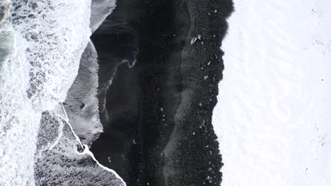 AERIAL:-Overhead-Birdsview-of-Person-walking-on-Black-Beach-with-white-arctic-snow-in-Iceland-in-Winter-Snow,-Ice,-Waves,-Water