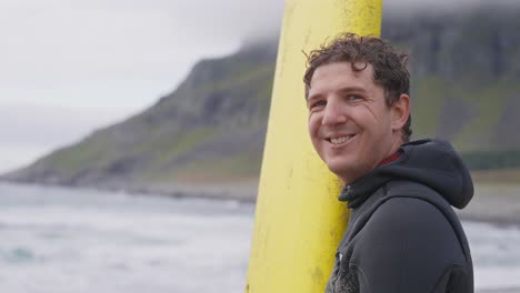 SLOW-MOTION-A-young-male-surfer-standing-next-to-his-yellow-beginner-surfboard-smiles-at-the-camera-then-stares-at-the-sea-in-Unstad-surf-beach,-Lofoten-Islands,-Norway