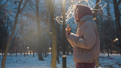 side view of young lady walking through winter park, enjoying snack, taking a bite from snack wrapped in paper, casual walking, winter evening with snow and park lights glowing in background