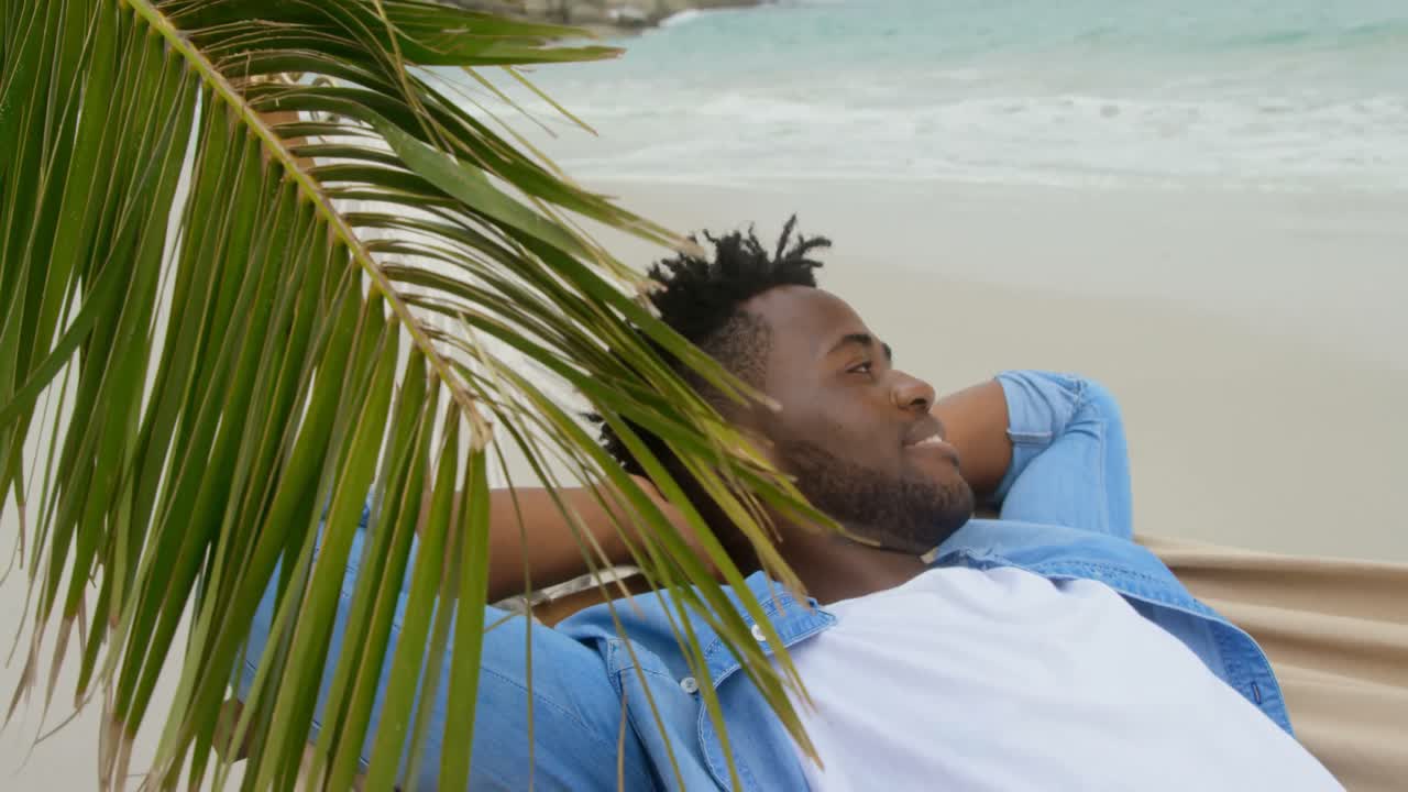 Side view of African american man relaxing in a hammock on the beach 4k