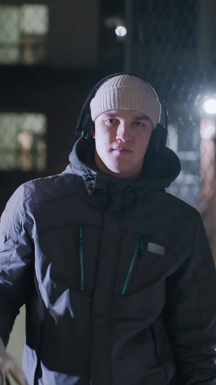 Tired man leaning on goal post, wearing gloves, beanie, and headphones under bright night lighting, focusing intently with urban lights reflecting in chain-link fence background