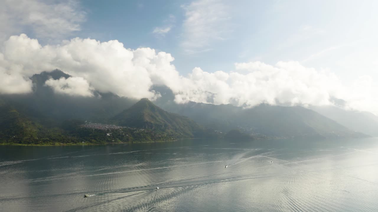 Boats drive by across calm Lake Atitlan Guatemala as clouds bunch on ridge line peaks