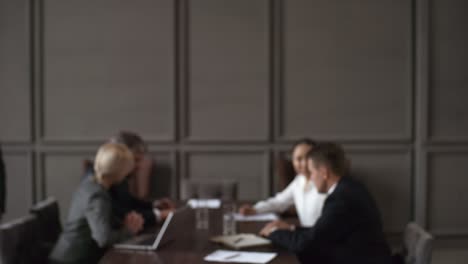 portrait of middle eastern businessman in suit entering frame, standing with crossed arms and looking at camera while in blurred background group of business executives sitting around table during meeting