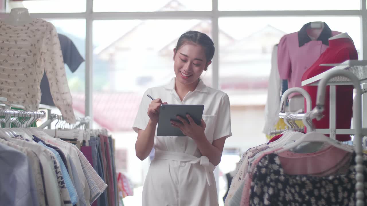 옷 가게의 아시아 여성 소유자가 태블릿에 메모를 쓰고 레일에서 재고를 확인하는 동안 카메라에 미소 짓고 있습니다.
