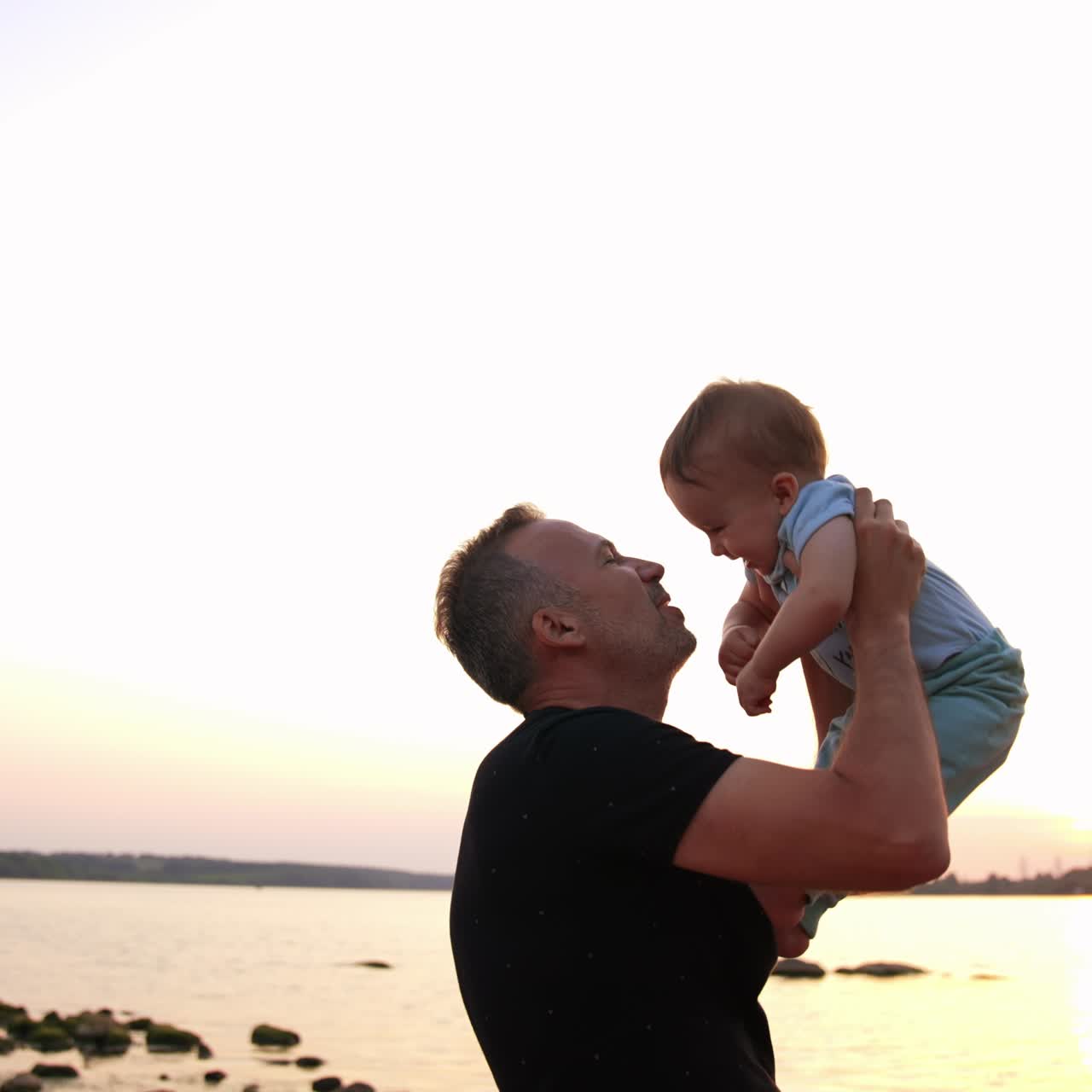 Loving dad talking to and kissing his little son. Laughing baby boy stepping his bare feet by father's chest. River at backdrop