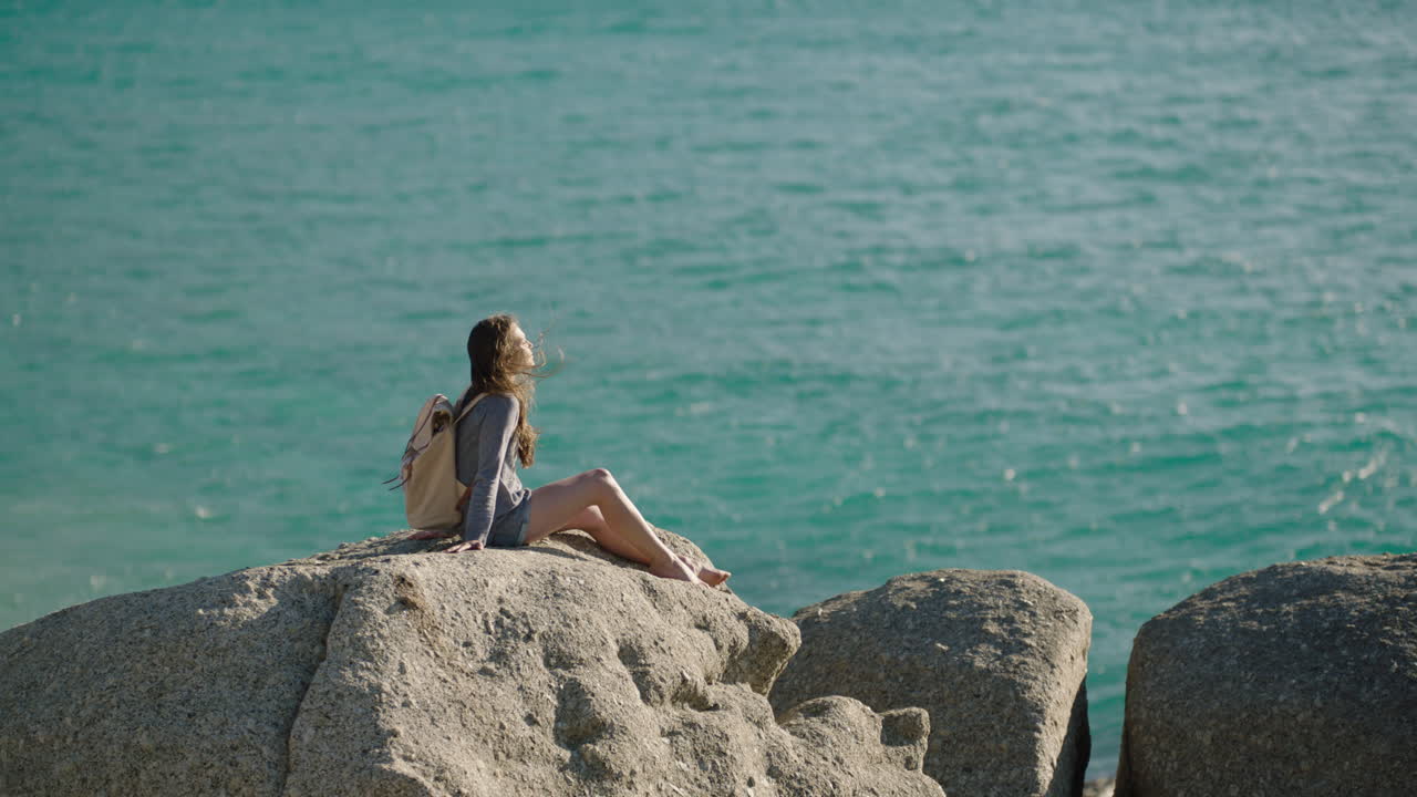 매력적인 젊은 독립적인 여성 관광객이 평화로운 무모한 휴가 라이프 스타일을 즐기고 바위 위에 앉아 아름다운 바다 해안 배경에서 휴식을 취하고 있습니다.