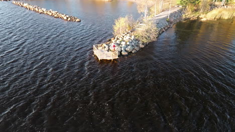 Fisherman-On-Wooden-Pier-With-Rocky-Lakeshore,-Black-Lake-Water-In-Sweden