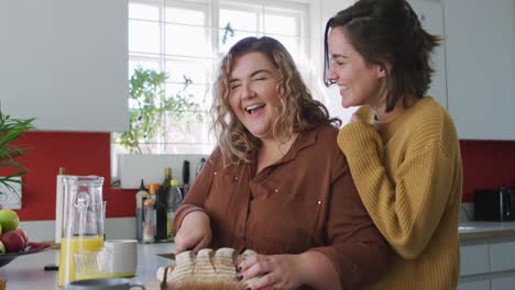 feliz pareja de lesbianas caucásicas cortando pan y riendo en la cocina