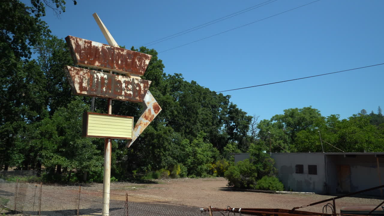 Rotating left pan shot of a rusty retro store sign out front of an abandoned lumber yard on a dirt road with mountains in the background on a bright sunny day