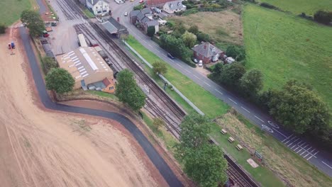 Aerial-tracking-backward-alongside-a-small-train-station-and-tracks,-with-a-busy-road-behind,-and-country-landscape-surrounding