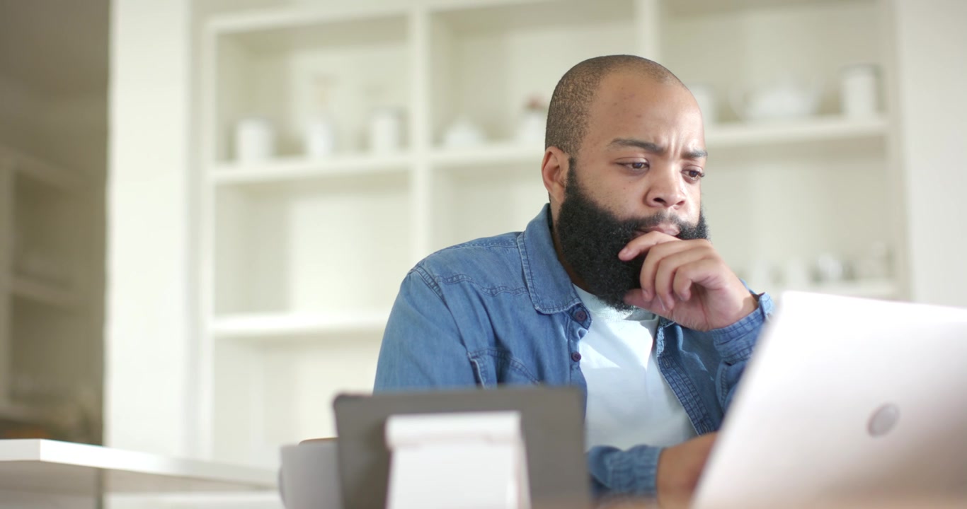 African American man in 30s typing on home laptop in denim shirt, analyzing screen, blurred stand