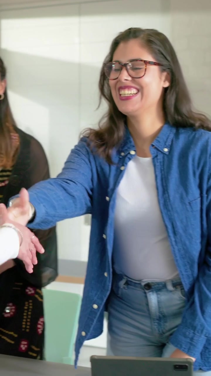 Video verticale: Donna che si alza in piedi per salutare un collega che entra in ufficio, stringendosi la mano, seduta vicino a un tablet per lavoro
