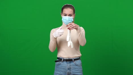 concentrated young woman in coronavirus face mask putting on protective gloves posing on green screen. portrait of young slim caucasian lady getting ready for going out on covid-19 pandemic outbreak