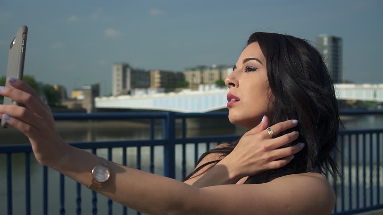 hermosa morena modelo hispana mira su teléfono y se toma selfies junto al río támesis en londres durante la brillante luz del día del verano, en reino unido