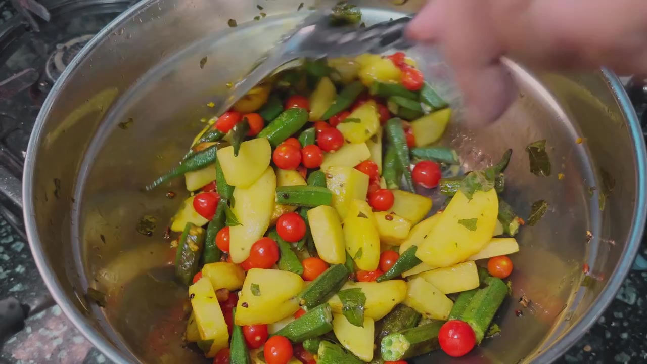 Vista aproximada de quiabo fresco, batatas e tomates cereja sendo mexidos dentro de uma panela de aço com especiarias e ervas visíveis durante o preparo caseiro de vegetais em um ambiente de cozinha