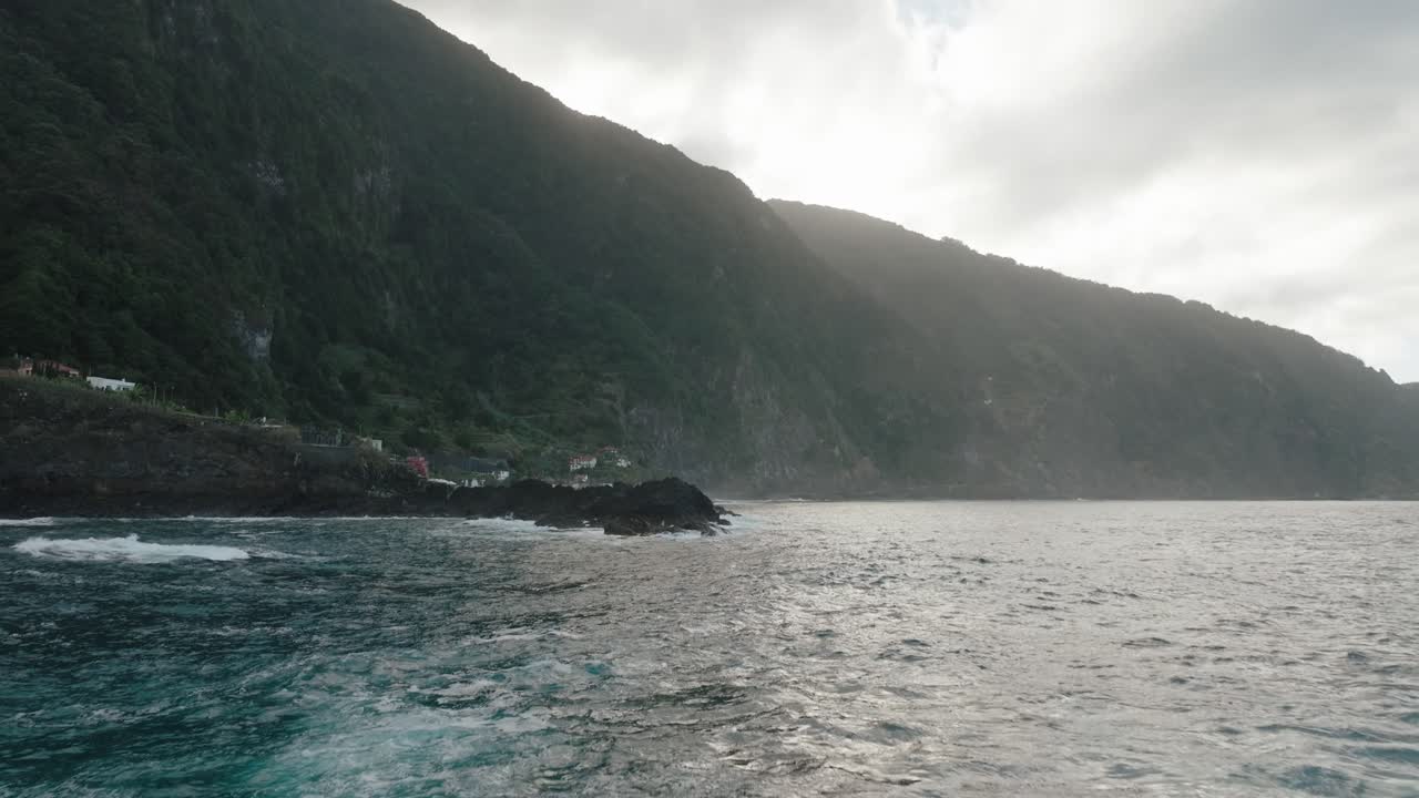 A man relaxes in a volcanic tide pool as waves crash beyond the rocks, facing the vast ocean and cliffs of Seixal, Madeira.