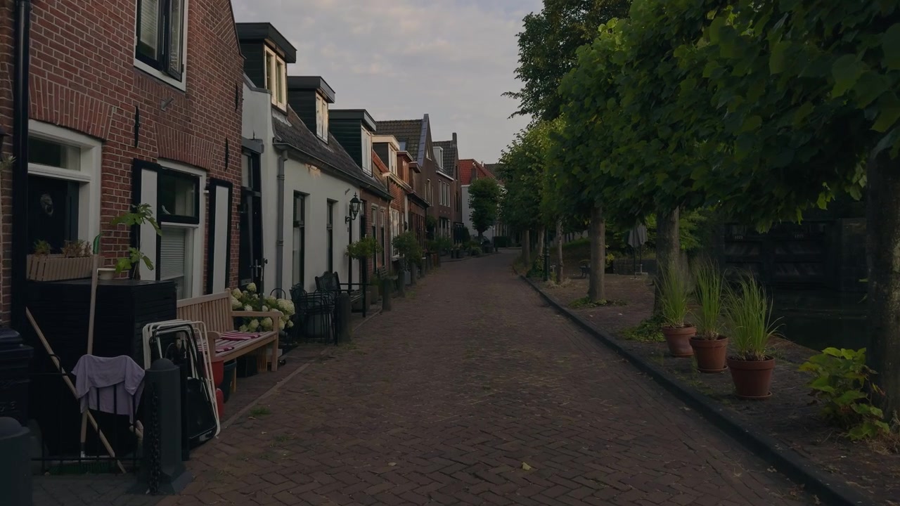POV, der går gennem en typisk hollandsk gade flankeret af charmerende kanalhuse og løvtræer langs vandet under en blød blå sommerhimmel.