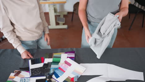 fashion creatives laying cloth on worktable for design project, adjusting soft gray fabric over colorful swatches and sewing patterns, preparing materials for creative garment production
