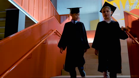 dos estudiantes de jardín de infantes con toga y birrete hablando juntos mientras bajan las escaleras en la ceremonia de graduación de preescolar